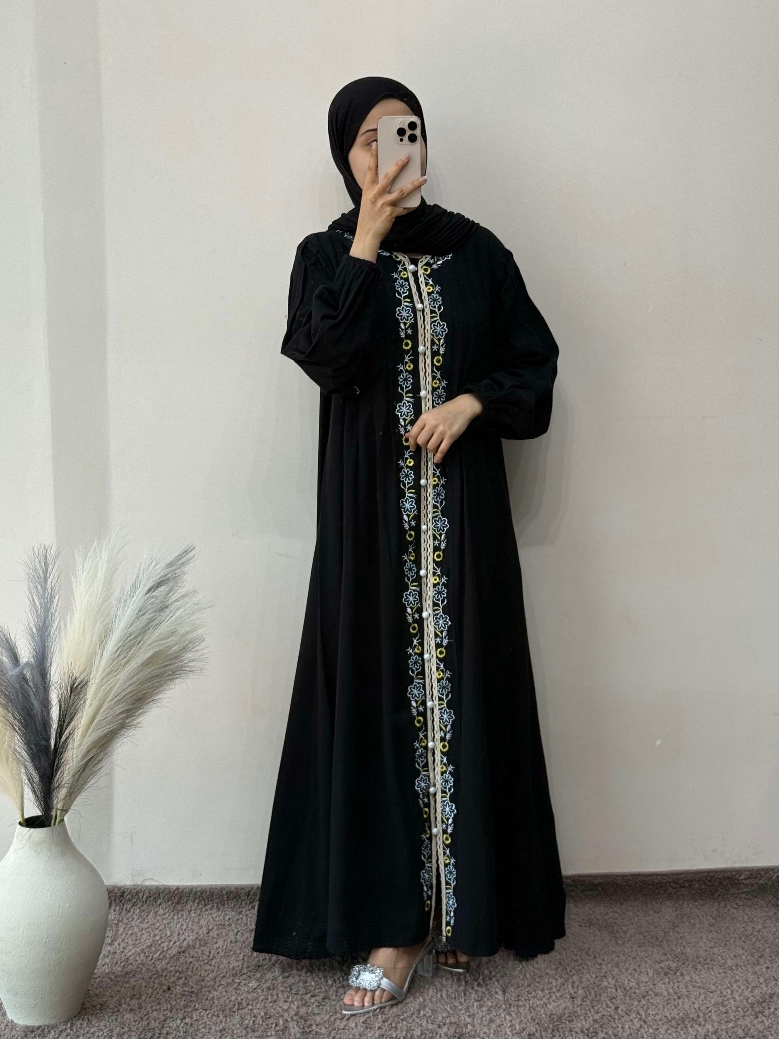 Person wearing a black abaya with gold embroidery in a minimal indoor setting.