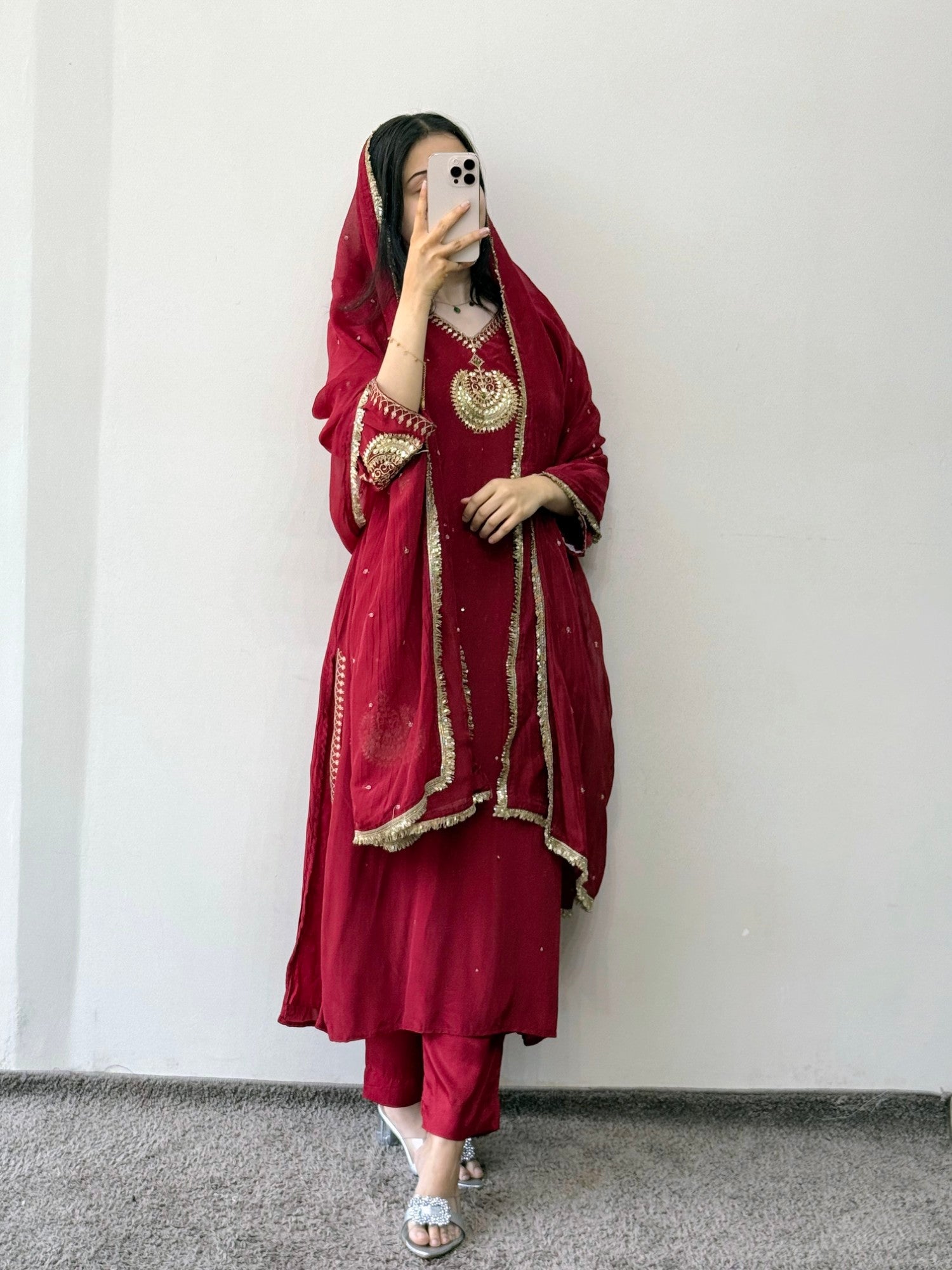 Person wearing a red traditional outfit with gold embroidery against a white wall.