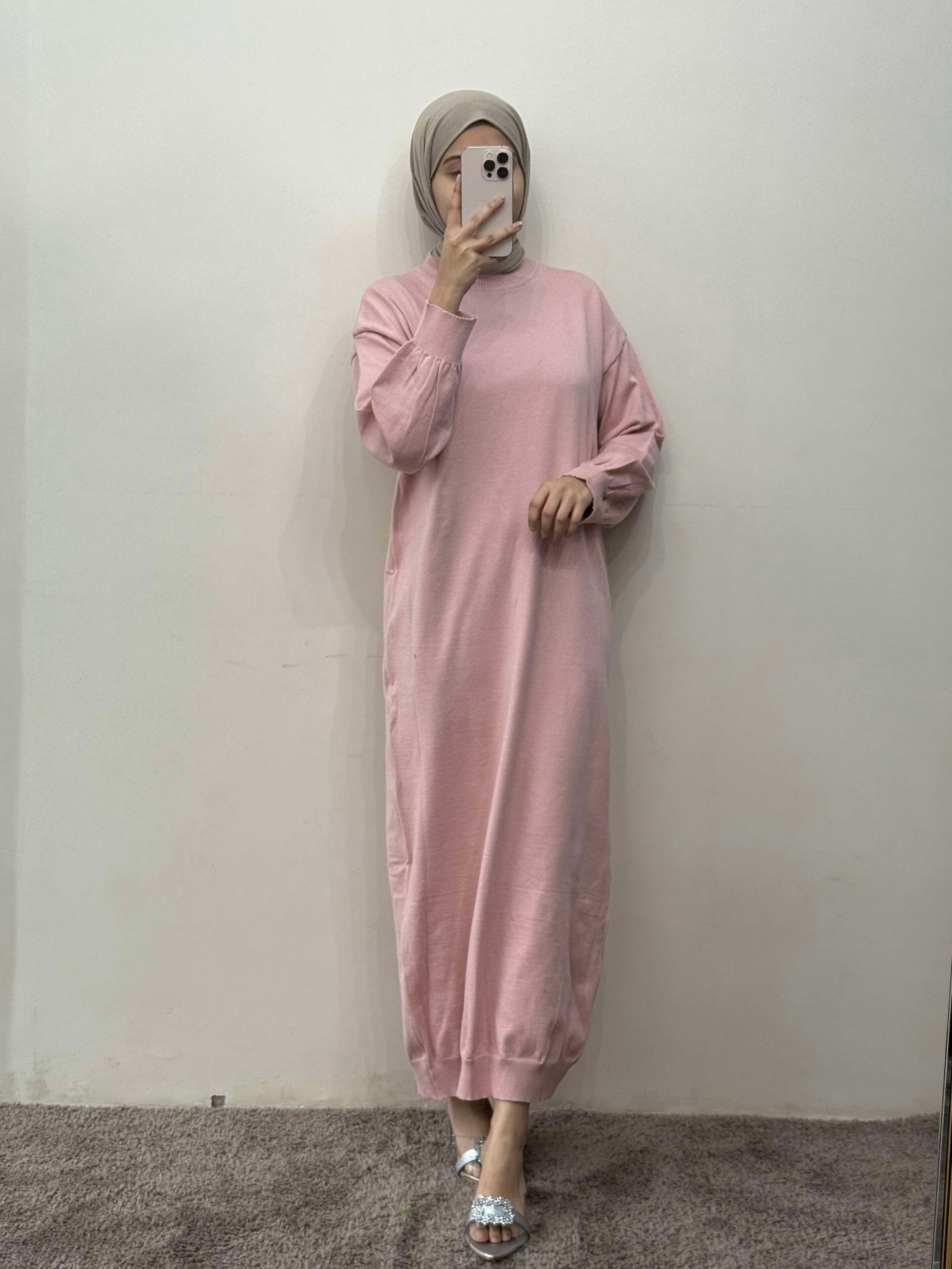Person wearing a pink dress and headscarf against a plain white wall.
