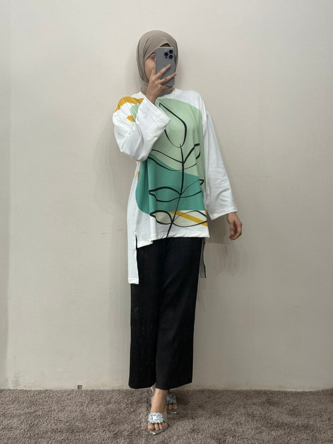 Person wearing a white shirt with a green abstract design and black pants against a plain wall.