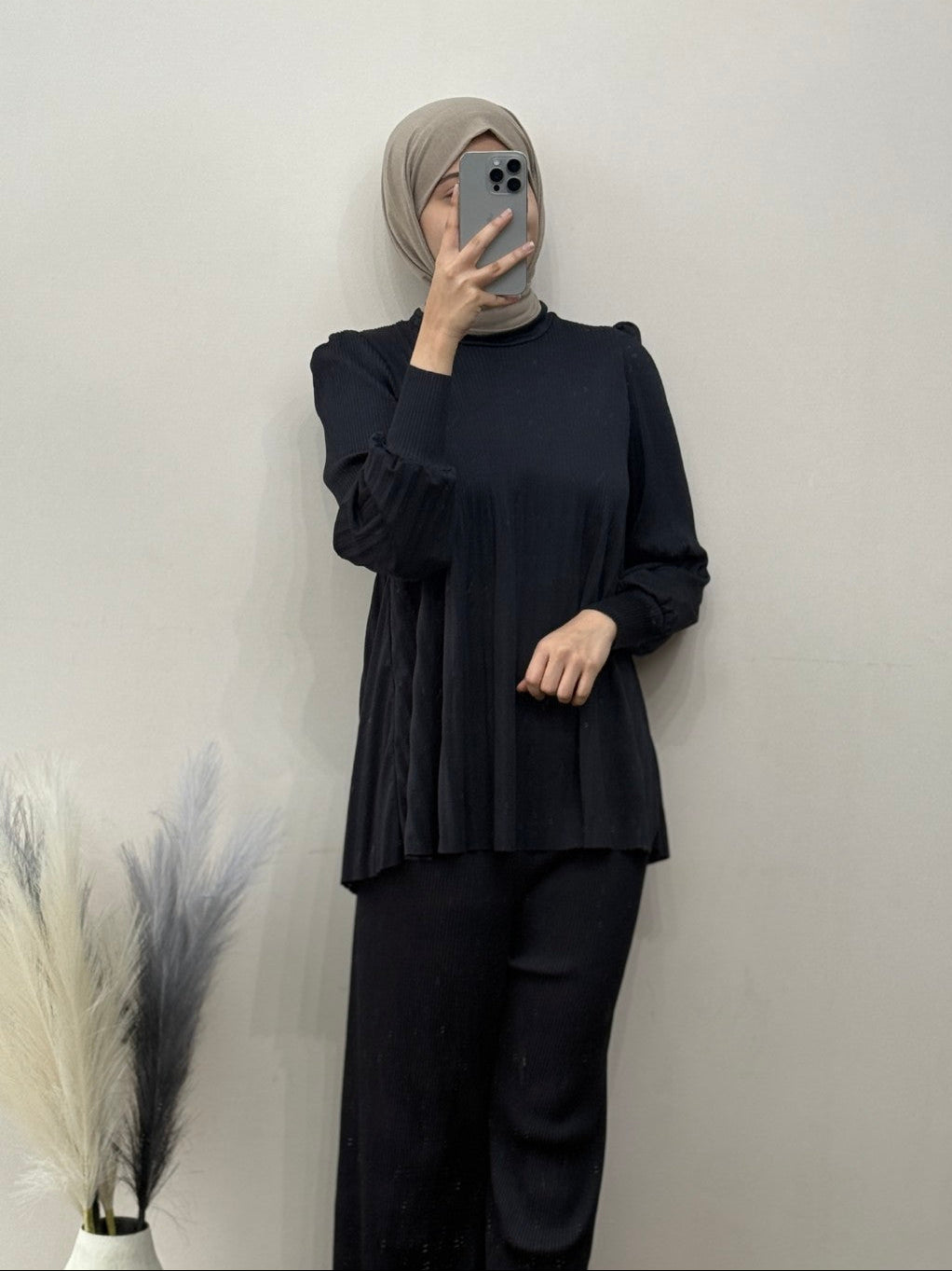 Person wearing a black outfit with a beige hijab against a plain background