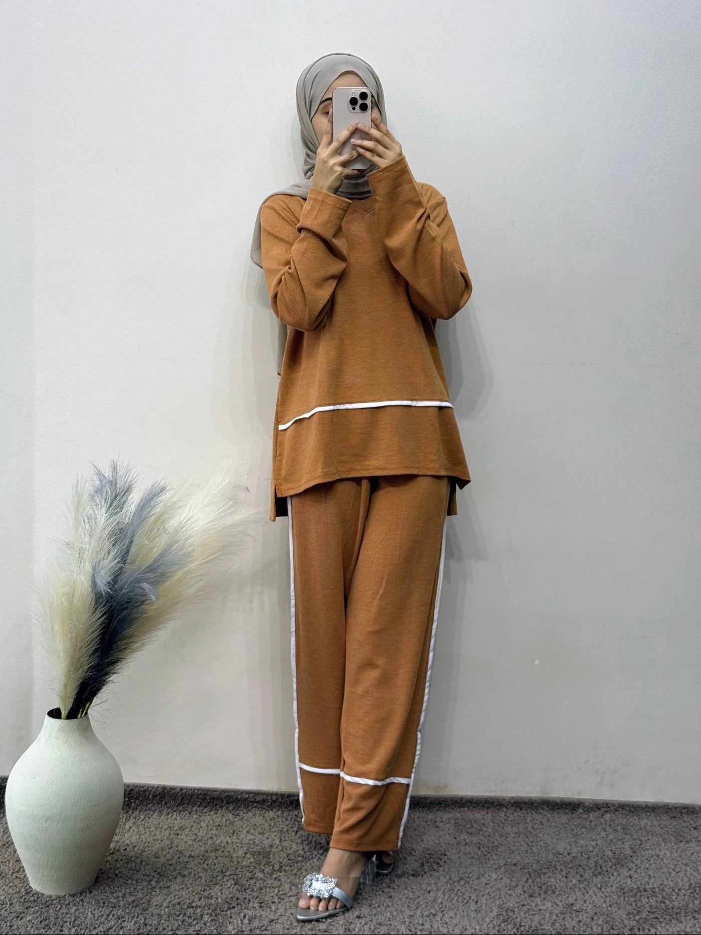 Person wearing a brown outfit with white trim, standing against a plain wall with a vase and plant decoration.