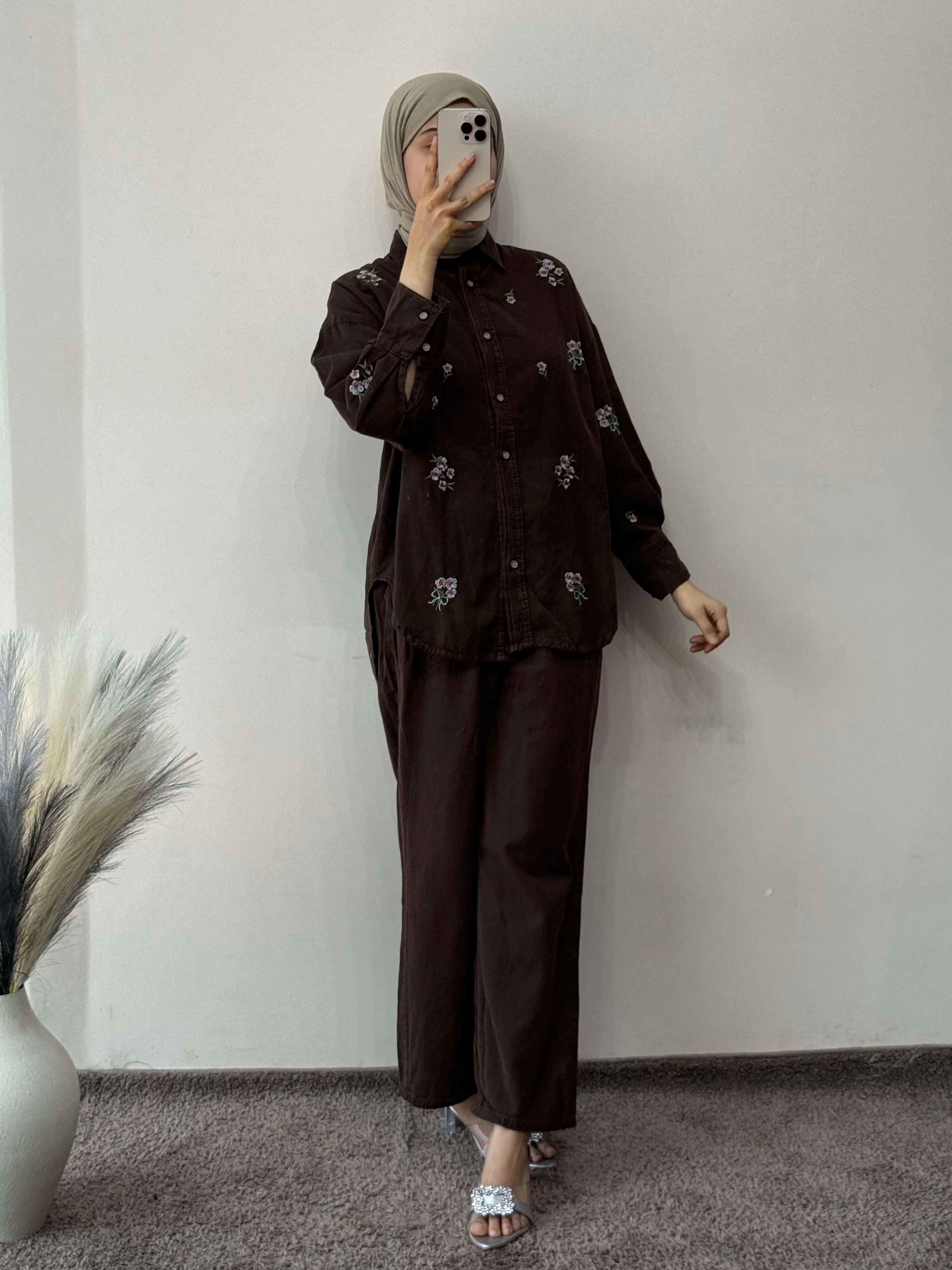 Person wearing a dark brown outfit with floral patterns against a plain wall.