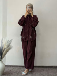 Person wearing a dark brown outfit standing against a plain wall with a vase of dried plants on the left.
