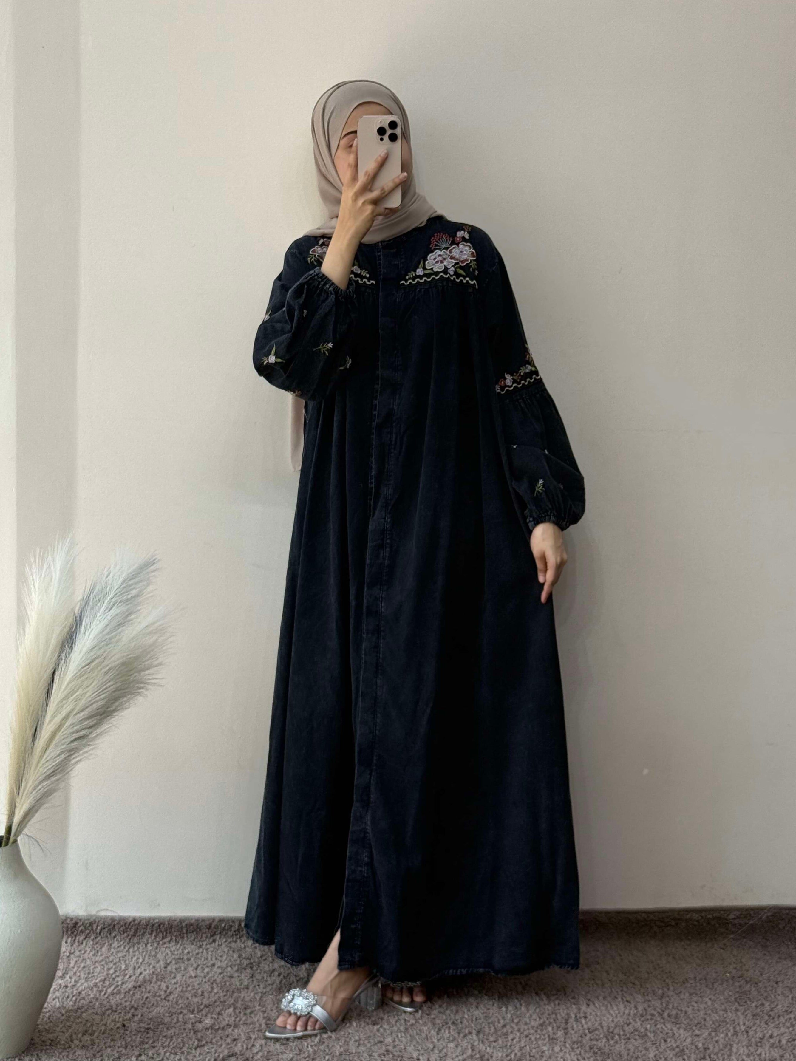 Person wearing a black dress with white embroidery against a plain wall.