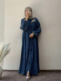 Person wearing a blue dress with floral embroidery against a plain wall.