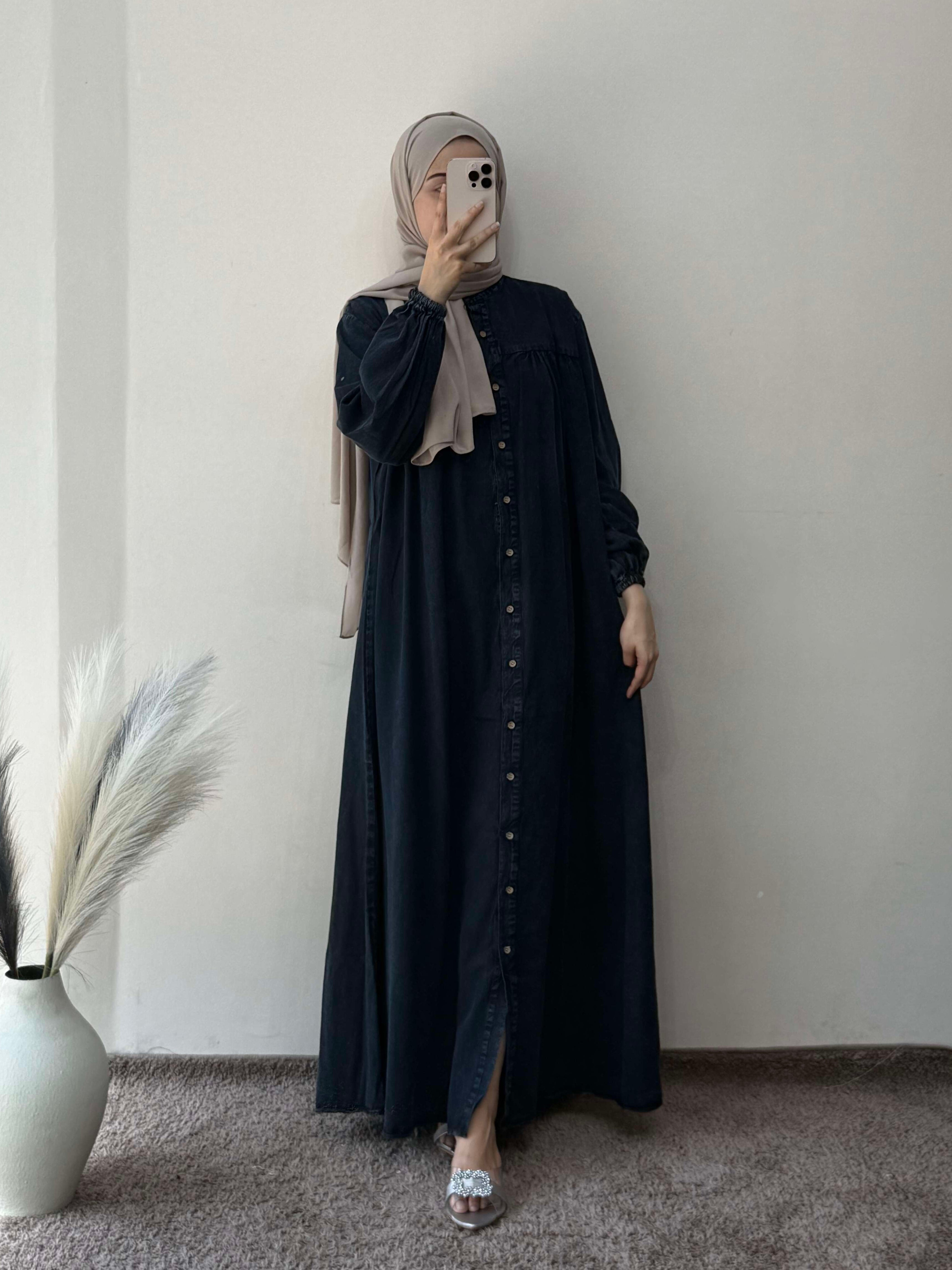 Person wearing a long black dress with a hijab in a room with a white wall and decorative elements.