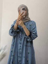 Person wearing a denim dress and hijab holding a phone against a plain background