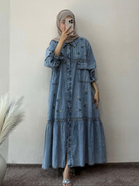 Person wearing a long denim dress standing against a plain wall with a vase of dried plants on the left.