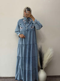 Person wearing a blue denim dress with floral embroidery in front of a plain wall.