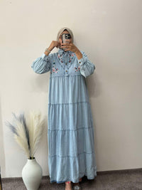 Person wearing a light blue dress with floral embroidery in front of a plain wall.