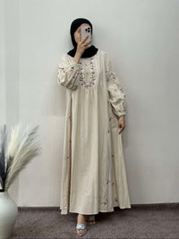 Person wearing a beige embroidered dress with a black hijab against a plain wall.