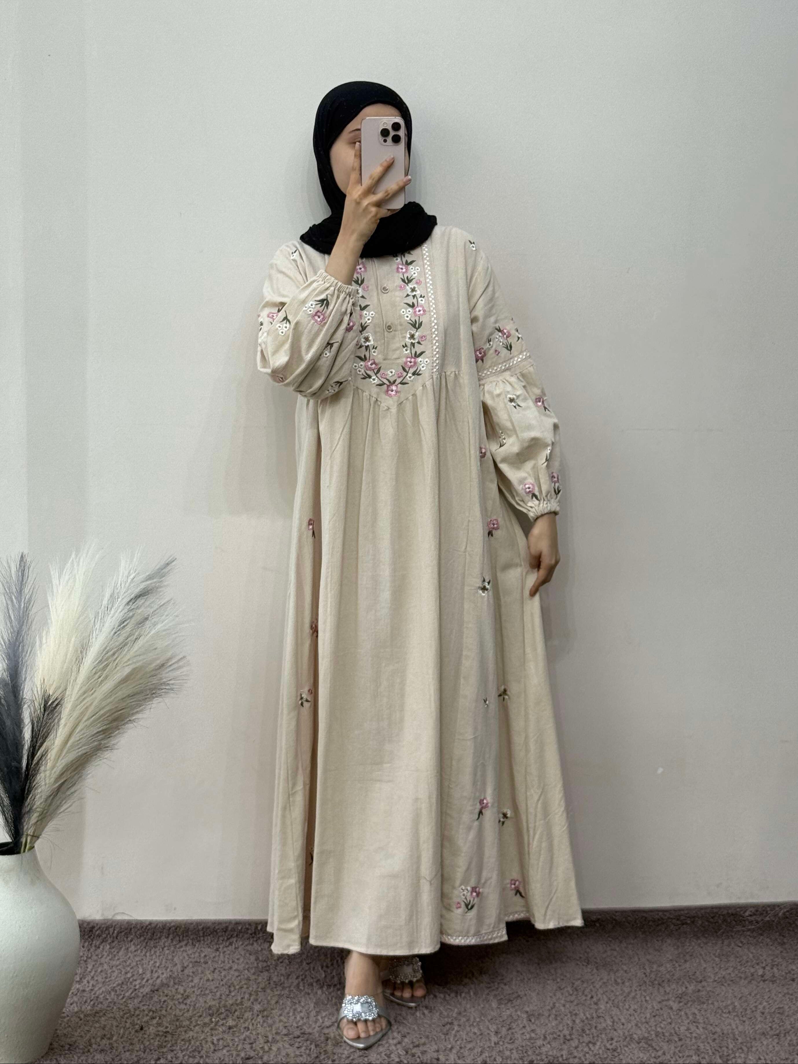 Person wearing a beige embroidered dress with a black hijab against a plain wall.