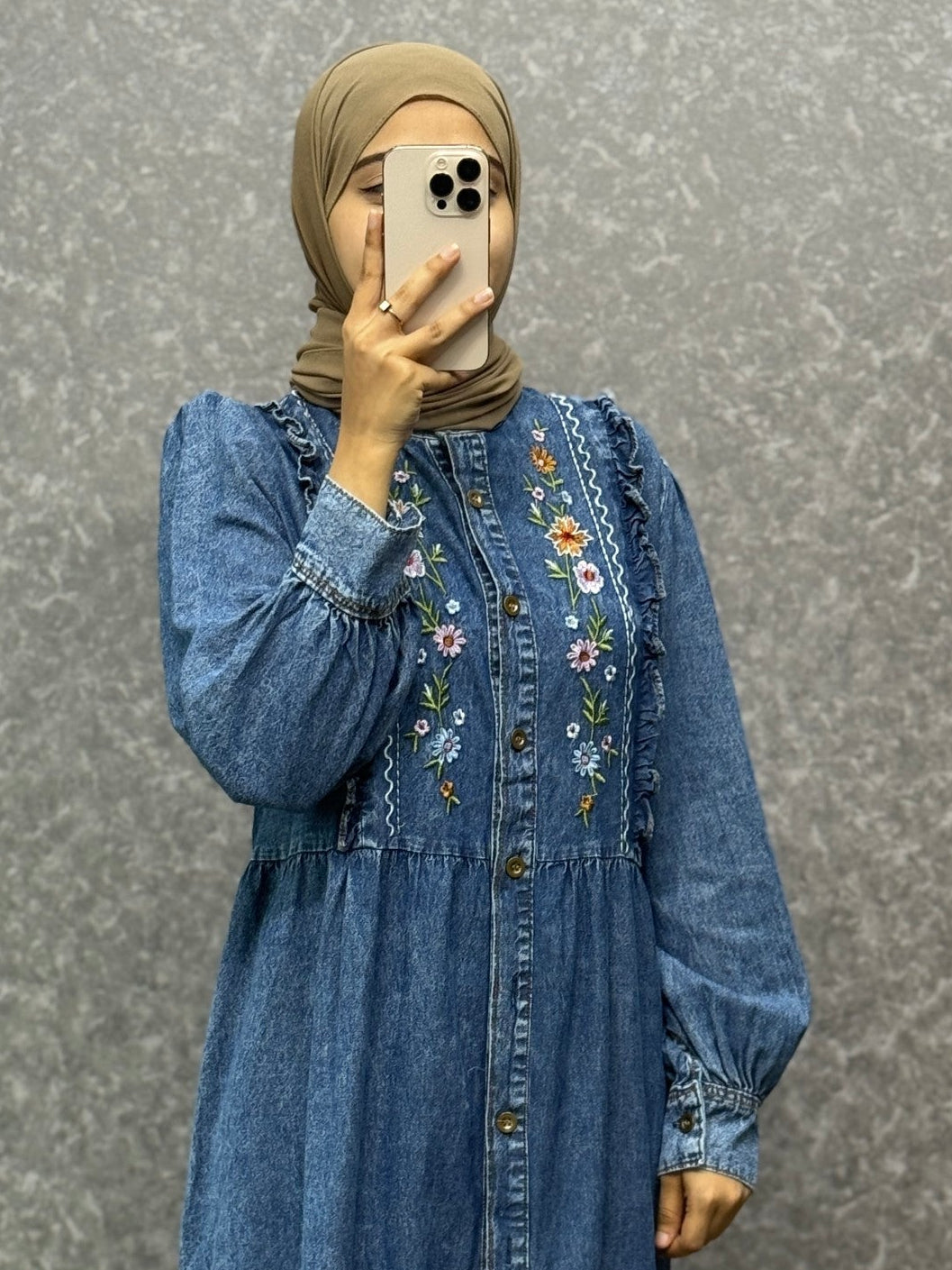 Person wearing a long blue denim dress with floral embroidery against a gray background