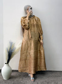 Woman wearing a long beige dress with a pattern, standing against a plain wall with a vase and plant decoration.