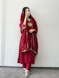 Person wearing a red traditional outfit with gold embroidery against a white wall.