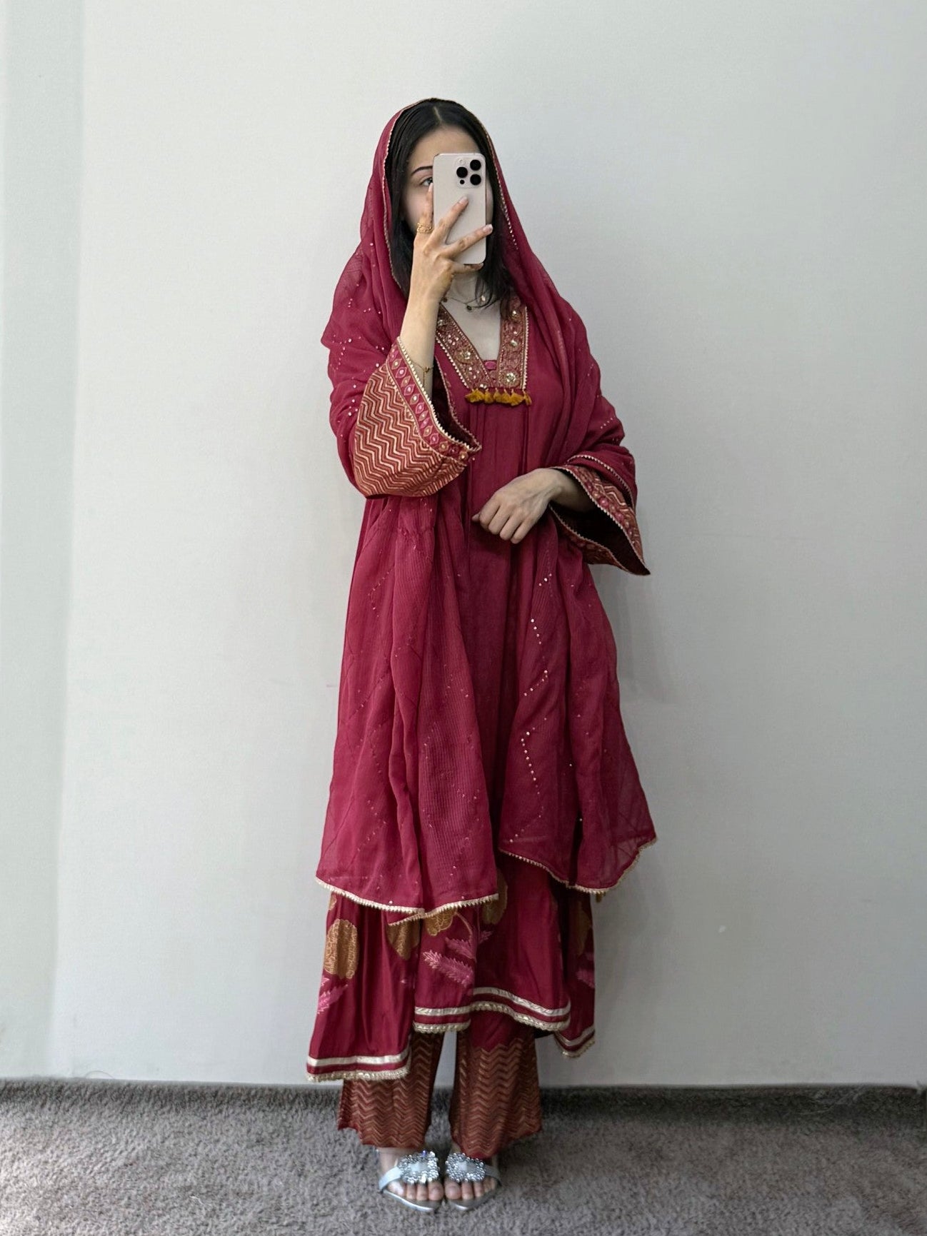 Person wearing a traditional maroon outfit with gold embroidery against a plain white wall.