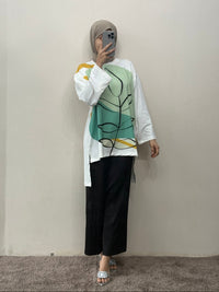 Person wearing a white shirt with a green abstract design and black pants against a plain wall.