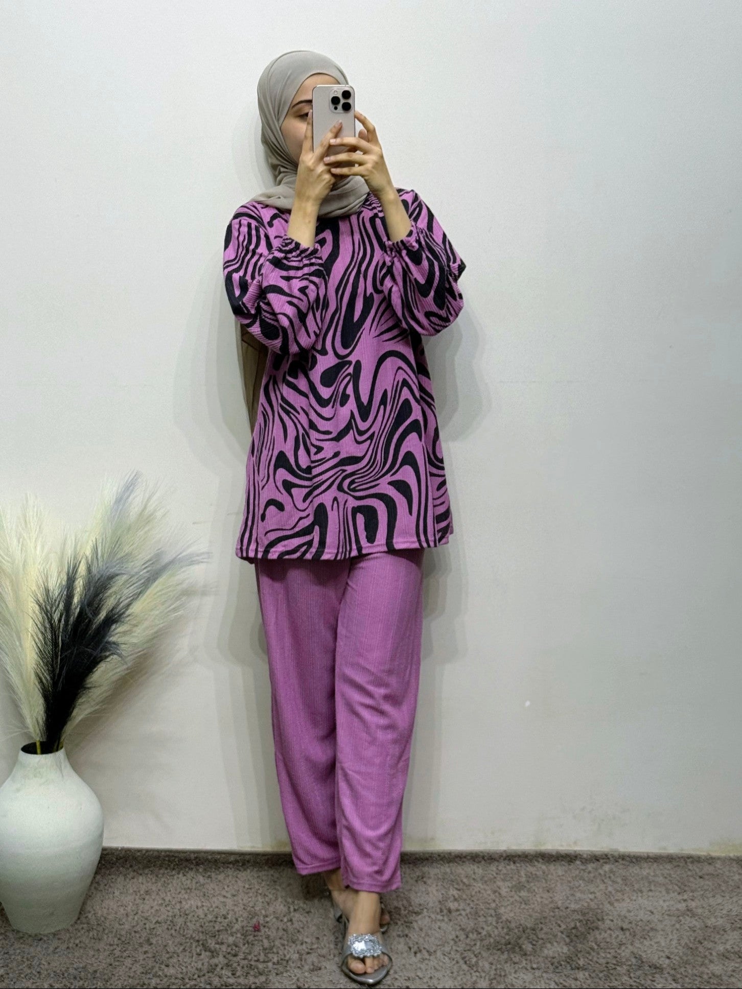 Person wearing a purple and black patterned top and matching pants, standing against a plain white wall.