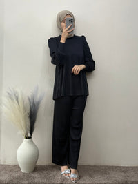 Person wearing a black outfit with a hijab, standing against a plain wall with a vase and decorative plants.