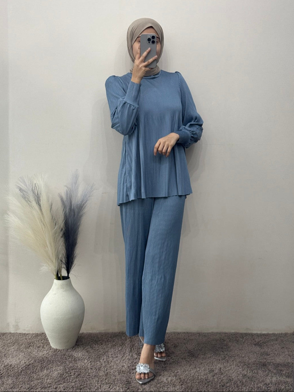 Person wearing a blue outfit with a hijab standing next to a white vase with decorative grasses on a gray carpet.