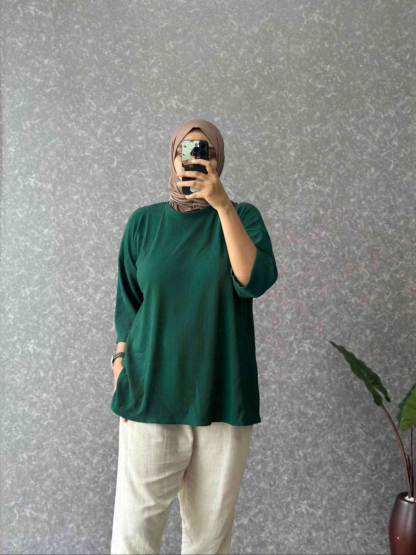 Person wearing a green top and white pants taking a mirror selfie against a gray wall.