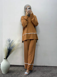 Person wearing a brown outfit with white trim, standing against a plain wall with a vase and plant decoration.