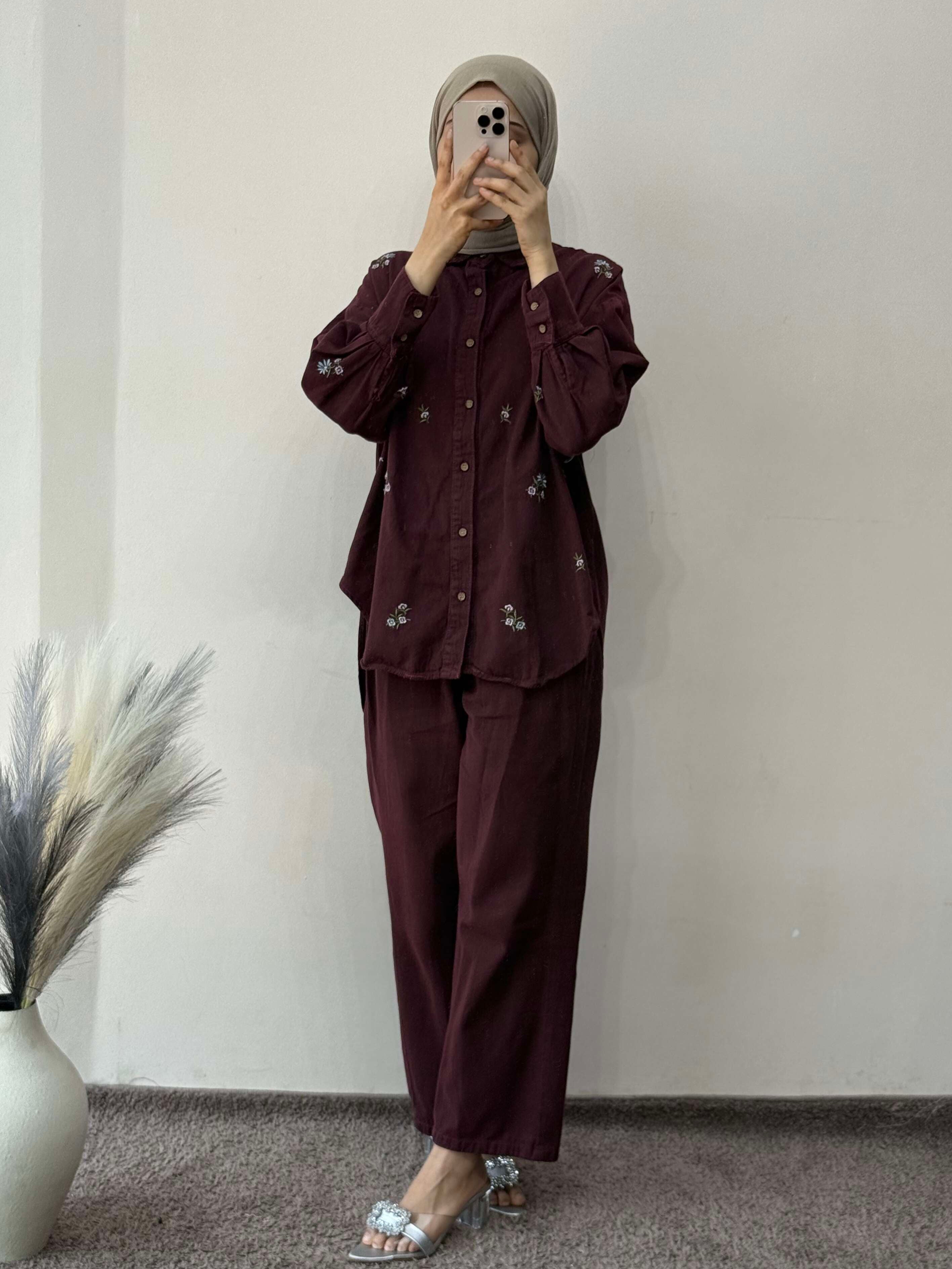 Person wearing a dark brown outfit standing against a plain wall with a vase of dried plants on the left.