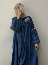 Person wearing a blue denim dress with floral embroidery against a plain background