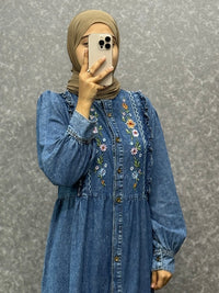 Person wearing a long blue denim dress with floral embroidery against a gray background