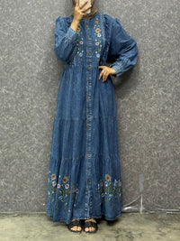 Person wearing a blue denim dress with floral embroidery against a gray wall.