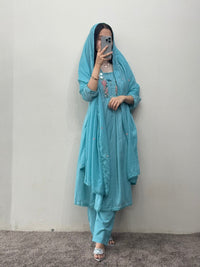 Person wearing a light blue traditional outfit with a matching headscarf against a plain background.