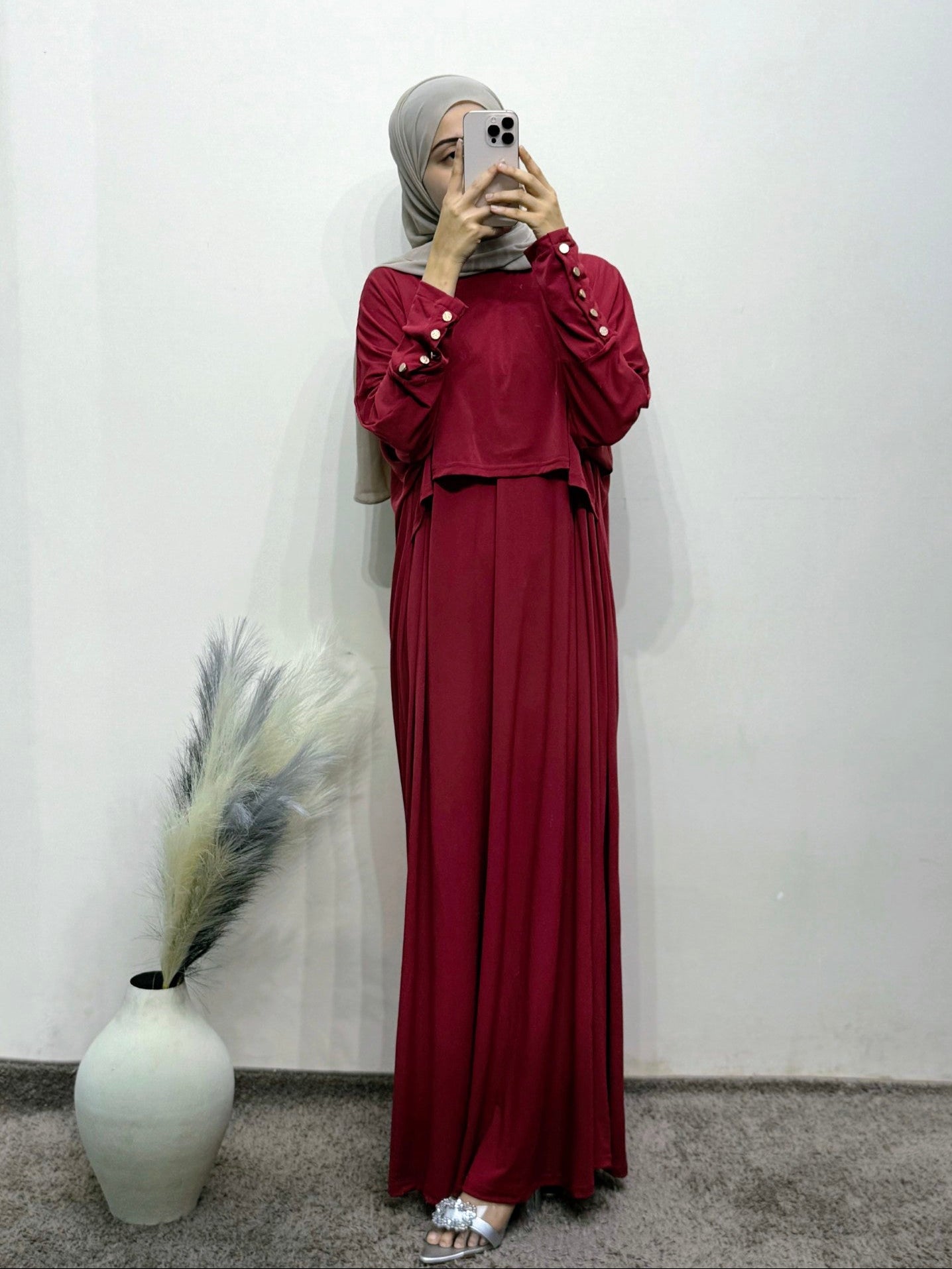 Person wearing a burgundy outfit with a hijab, standing against a white wall with a vase and dried plants.