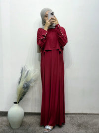 Person wearing a burgundy outfit with a hijab, standing against a white wall with a vase and dried plants.