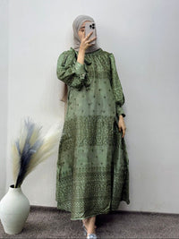 Person wearing a green patterned dress standing against a white wall with a vase and decorative plant.