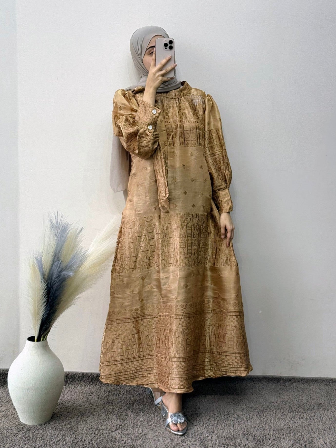 Woman wearing a long beige dress with a pattern, standing against a plain wall with a vase and plant decoration.