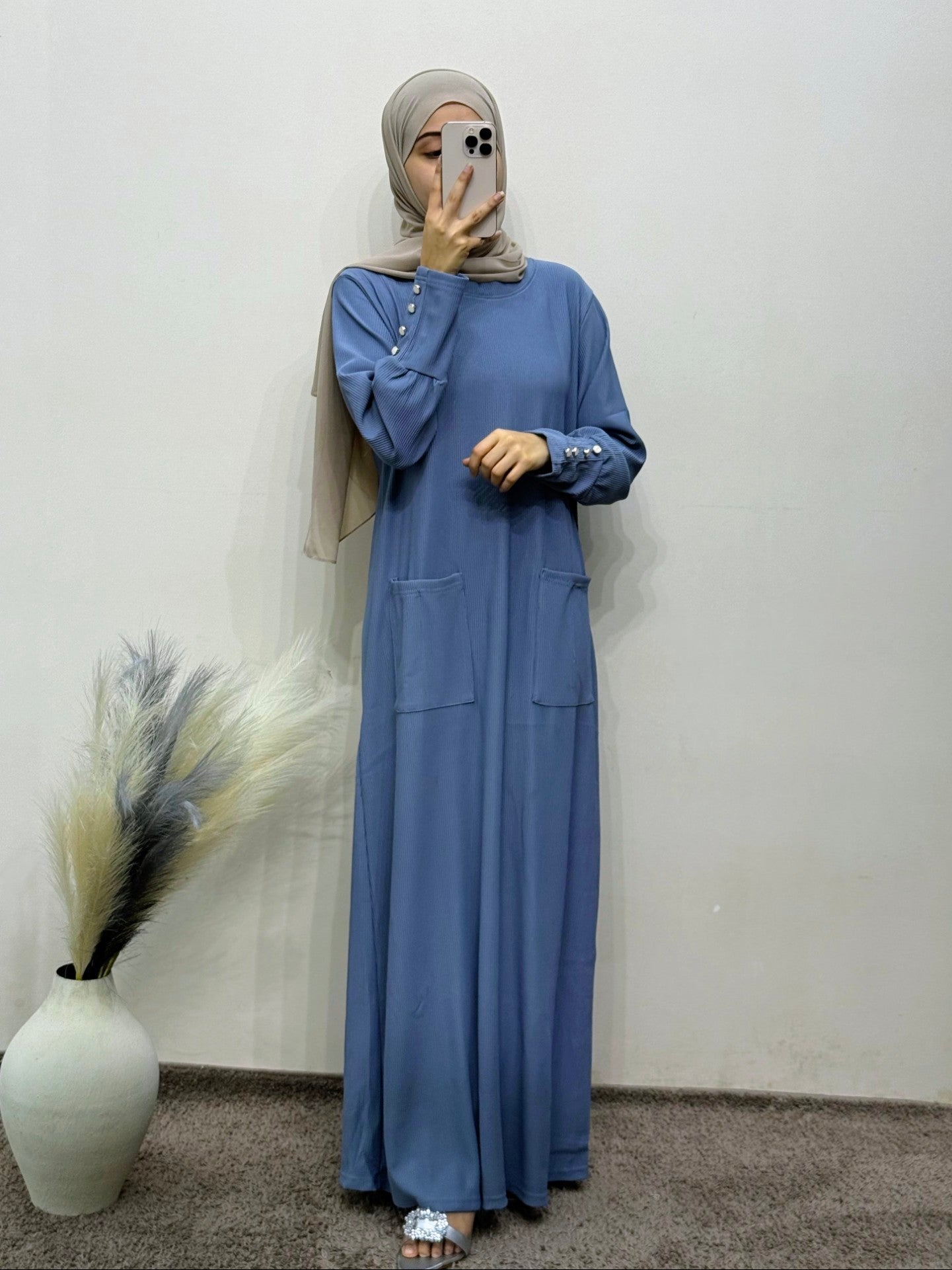 Person wearing a blue dress and hijab standing against a plain wall with a decorative vase.